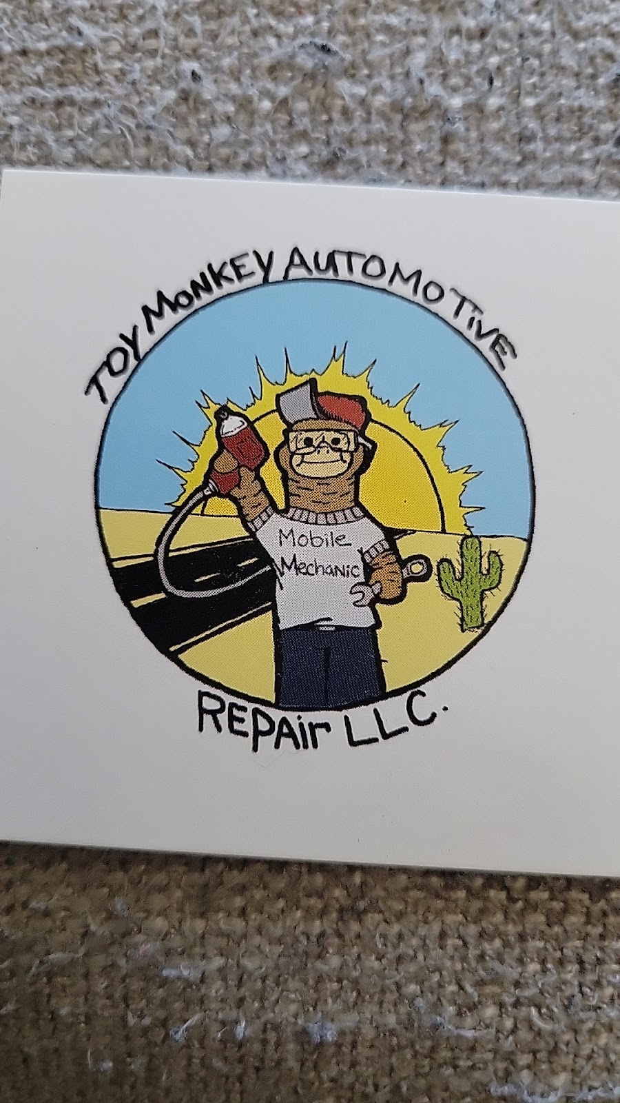 Hand-drawn Toy Monkey badge: a cartoon monkey in a Mobile Mechanic tee holding up a drill, with a desert sun, road, and cactus behind him